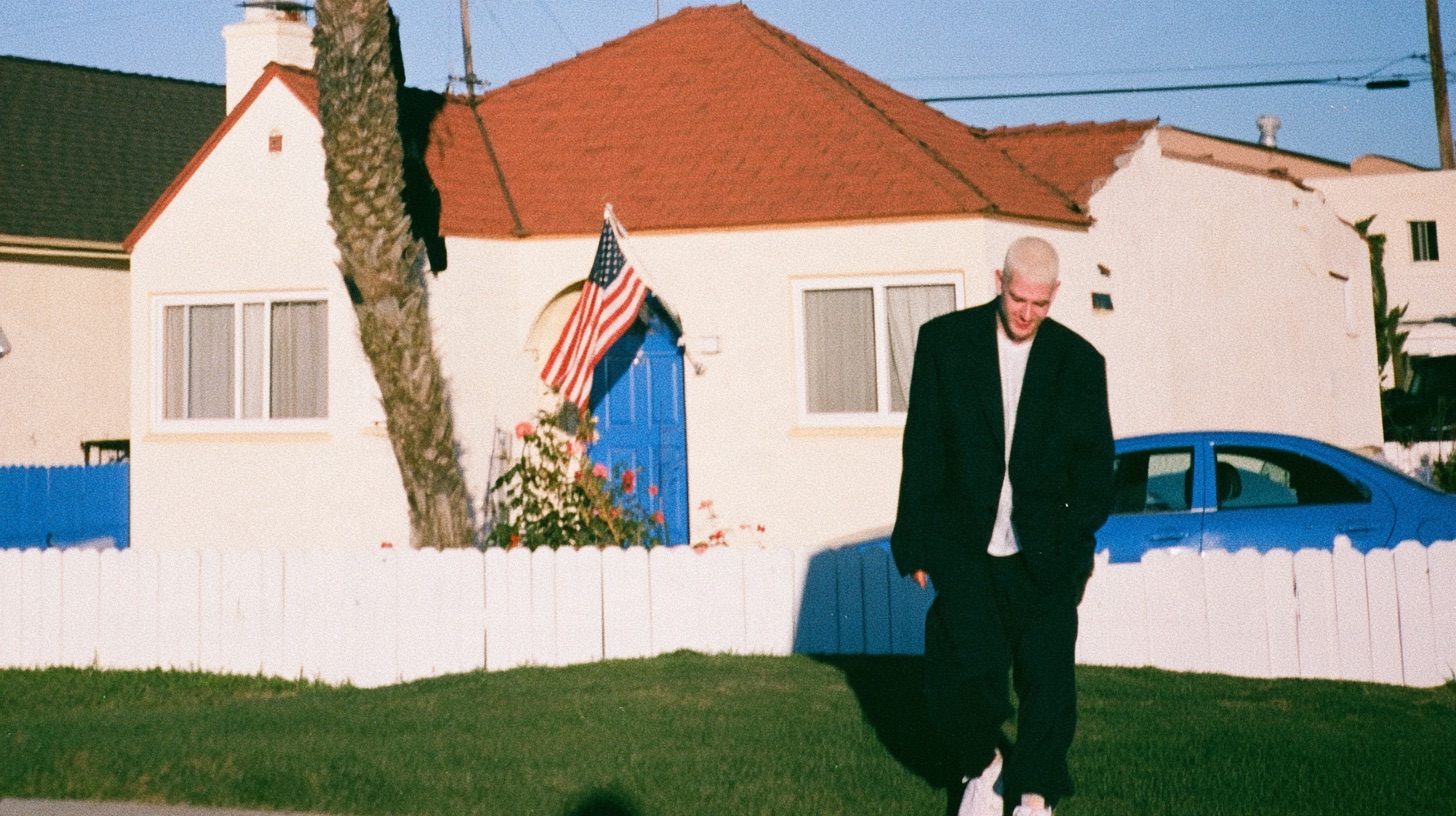 An adult standing outside a modest childhood house, looking back at it, late afternoon, quiet not sentimental