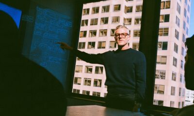 A charismatic tech CEO in a minimalist dark sweater stands at the head of a concrete table pointing to a procurement workflow chart sketched on a glass wall