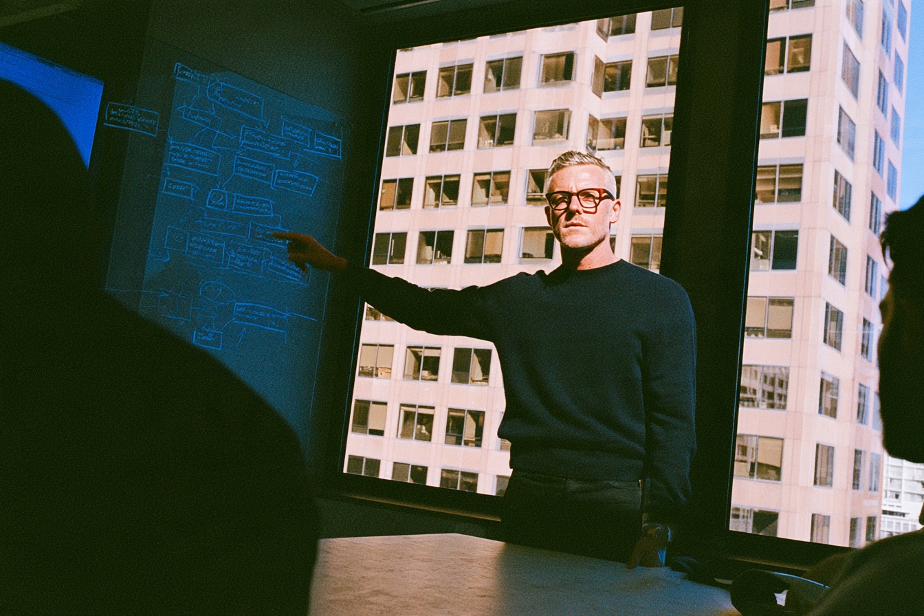 A charismatic tech CEO in a minimalist dark sweater stands at the head of a concrete table pointing to a procurement workflow chart sketched on a glass wall