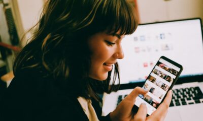 A female entrepreneur with a focused look and smile on her face using a smartphone to browse social media in front of a laptop