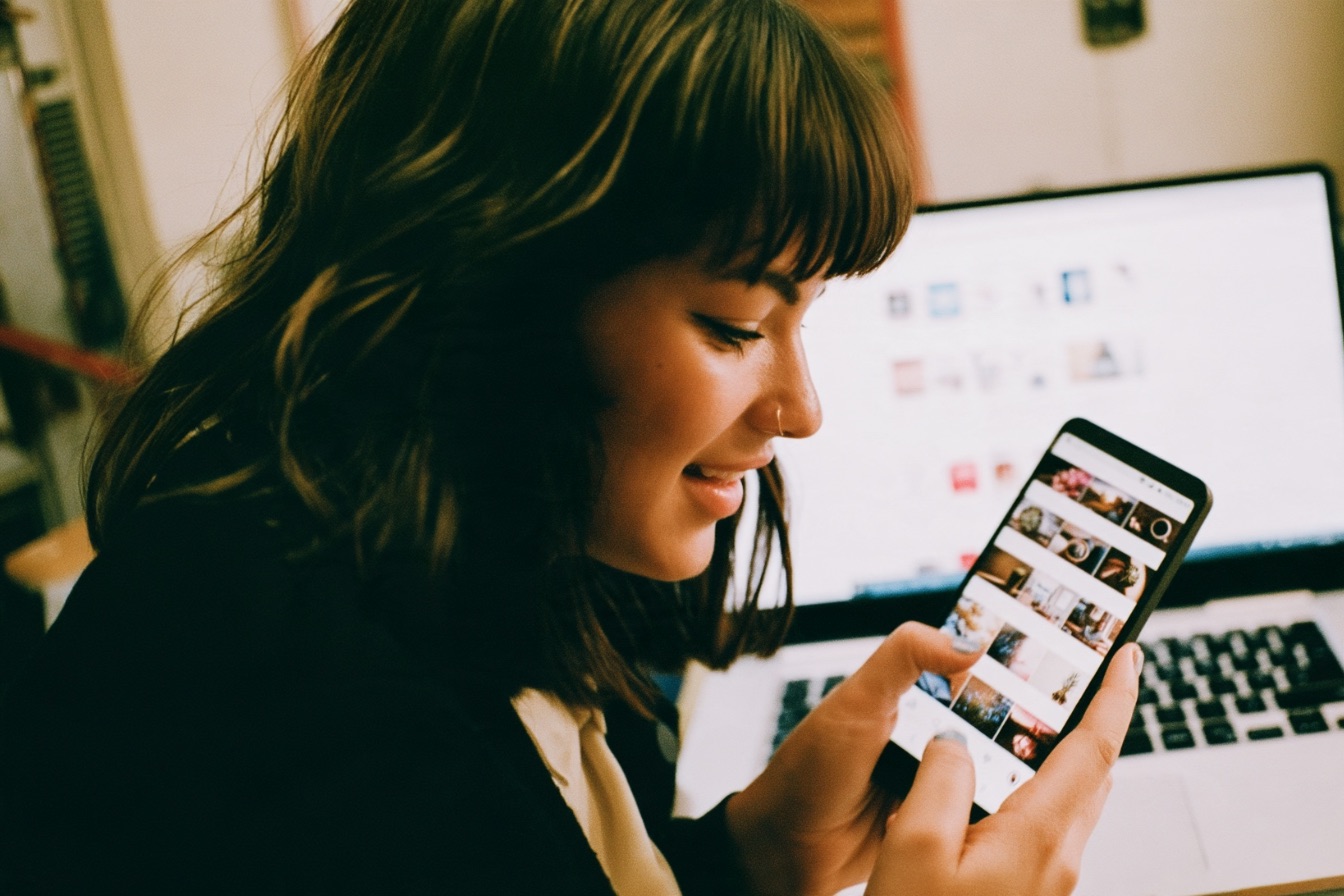 A female entrepreneur with a focused look and smile on her face using a smartphone to browse social media in front of a laptop
