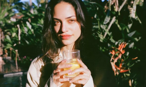A female founder in a minimalist cream linen robe standing in a sun drenched architectural kitchen holding a clear glass of warm lemon water with both hands
