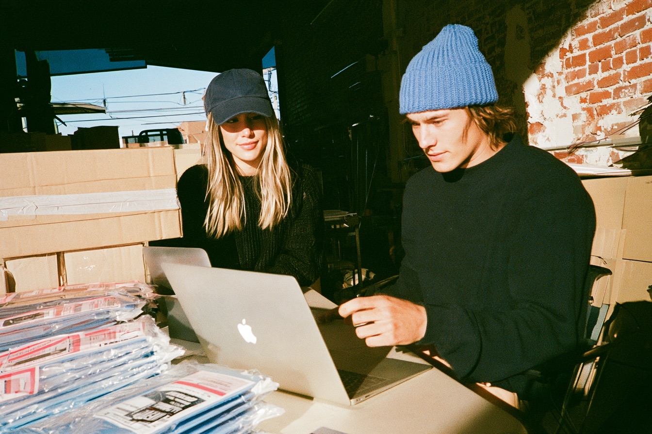A man and woman entrepreneur looking at a laptop screen running an ecommerce store with products shipping out in the background