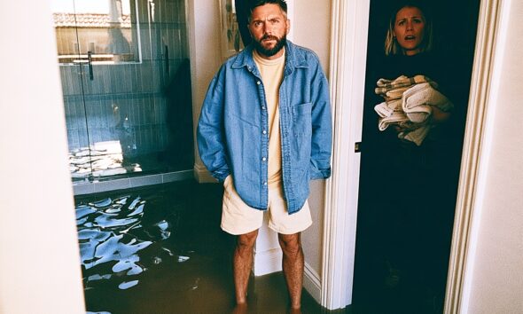 A man standing in water as his plumbing needs to be fixed in his shower and his wife is shocked