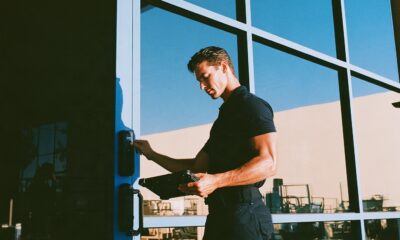 A rugged male security consultant in a fitted black polo shirt and tactical pants confidently pressing his thumb against a sleek matte black biometric smart lock on a modern glass front door