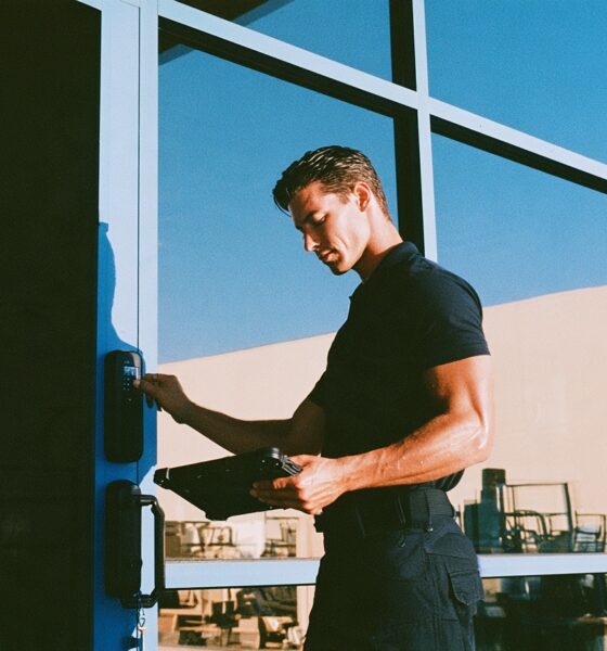 A rugged male security consultant in a fitted black polo shirt and tactical pants confidently pressing his thumb against a sleek matte black biometric smart lock on a modern glass front door