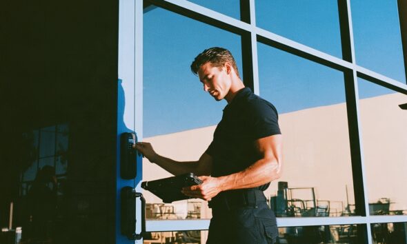 A rugged male security consultant in a fitted black polo shirt and tactical pants confidently pressing his thumb against a sleek matte black biometric smart lock on a modern glass front door