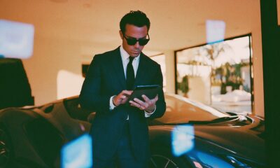 A sharp male entrepreneur in a tailored charcoal suit standing beside a sleek modern luxury sports car inside a sunlit glass architectural villa