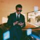 A sharp male entrepreneur in a tailored charcoal suit standing beside a sleek modern luxury sports car inside a sunlit glass architectural villa
