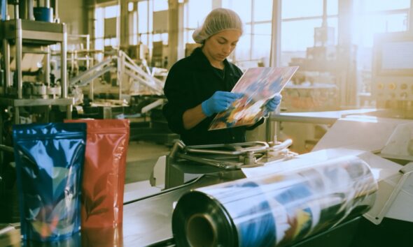 a focused female quality control worker in a hairnet and blue industrial gloves inspecting a sheet of brightly colored custom flexible packaging on a brightly lit factory floor Large