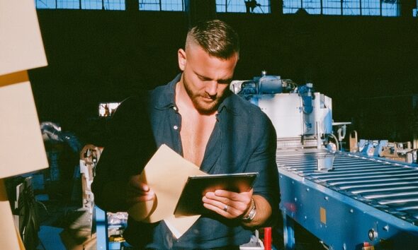 a male operations director standing on the floor of a massive industrial manufacturing facility holding a digital tablet and inspecting an automated conveyor belt line