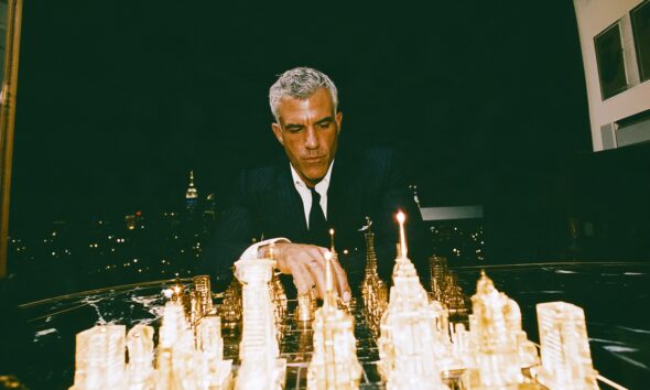 a sharply dressed male entrepreneur in his early forties sitting alone across an enormous luxury black marble chessboard inside a dark executive penthouse