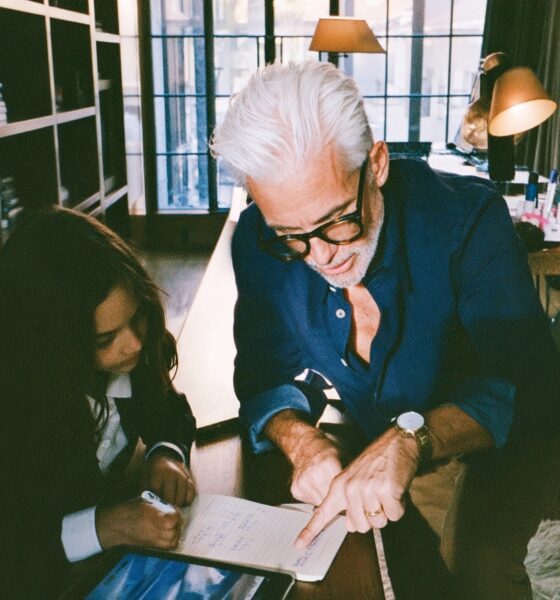 a sharply dressed tech billionaire founder father in a navy overshirt kneeling beside his ten year old daughter as she works through a math lesson on a digital tablet