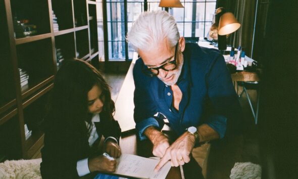 a sharply dressed tech billionaire founder father in a navy overshirt kneeling beside his ten year old daughter as she works through a math lesson on a digital tablet