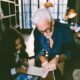 a sharply dressed tech billionaire founder father in a navy overshirt kneeling beside his ten year old daughter as she works through a math lesson on a digital tablet