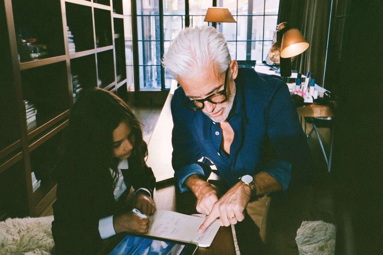 a sharply dressed tech billionaire founder father in a navy overshirt kneeling beside his ten year old daughter as she works through a math lesson on a digital tablet