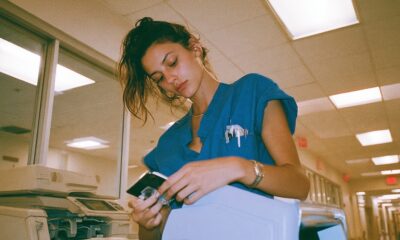 an exhausted nurse in teal scrubs holding a bulky physical medical CD in a sterile brightly lit hospital hallway and there is an old fax machine near her