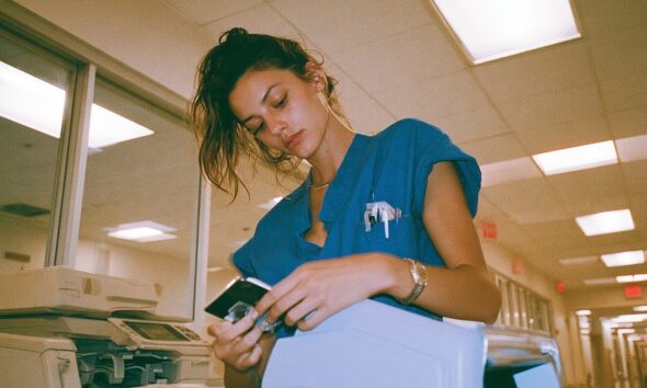 an exhausted nurse in teal scrubs holding a bulky physical medical CD in a sterile brightly lit hospital hallway and there is an old fax machine near her