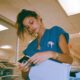 an exhausted nurse in teal scrubs holding a bulky physical medical CD in a sterile brightly lit hospital hallway and there is an old fax machine near her