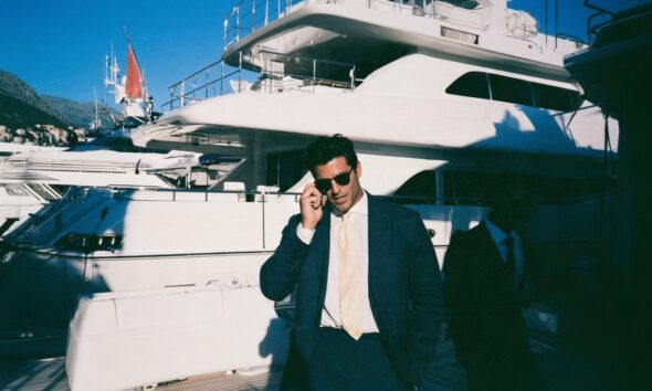 male entrepreneur in a navy linen suit standing confidently on the teak deck of a massive superyacht moored in a luxury marina in Montenegro
