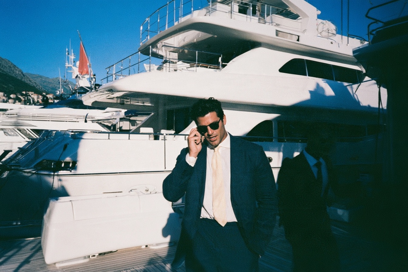 male entrepreneur in a navy linen suit standing confidently on the teak deck of a massive superyacht moored in a luxury marina in Montenegro