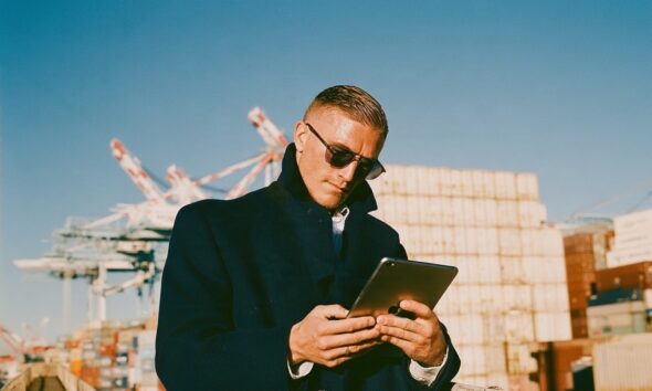 male logistics director in a tailored navy topcoat and dark wireframe aviators, with a digital apple tablet standing on the elevated catwalk of an international shipping port at dawn