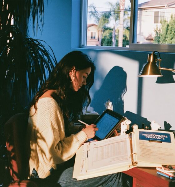 woman in a cream knit sweater updating a remittance ledger spreadsheet on a tablet while a homeowners insurance binder sits open