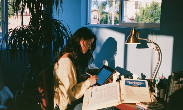 woman in a cream knit sweater updating a remittance ledger spreadsheet on a tablet while a homeowners insurance binder sits open
