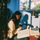 woman in a cream knit sweater updating a remittance ledger spreadsheet on a tablet while a homeowners insurance binder sits open