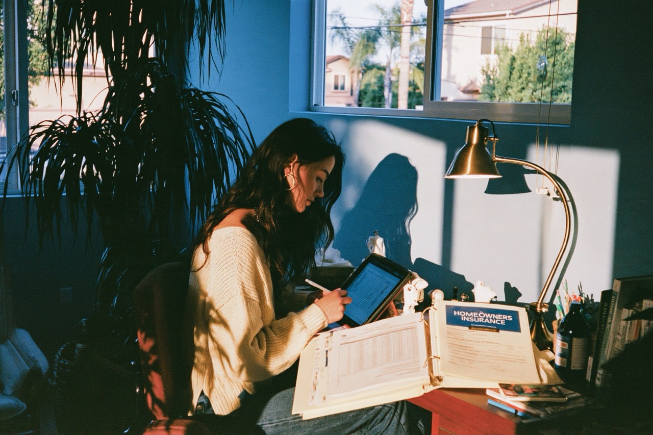woman in a cream knit sweater updating a remittance ledger spreadsheet on a tablet while a homeowners insurance binder sits open